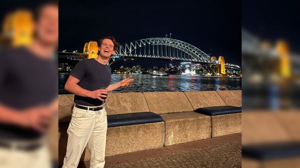 Will Barker stands outside at night, pointing at the Sydney Harbour Bridge in the background.