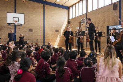 Musicians holding instruments perform in front of a group of seated children