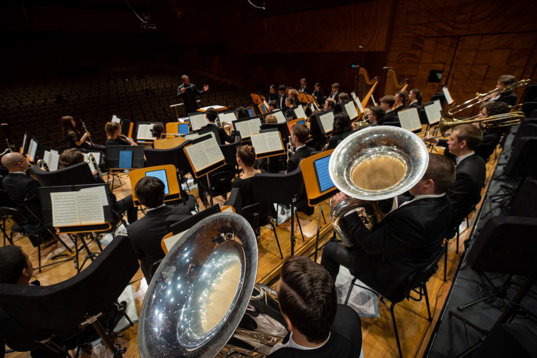 Students from the University of Melbourne Wind Symphony hold brass wind instruments and look out from the stage to the Hamer Hall seats in front of them.