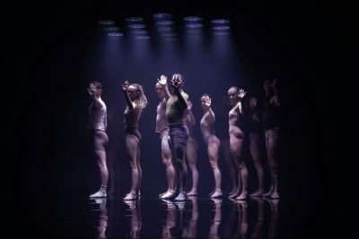 A group of dancers stand in profile on a darkened stage