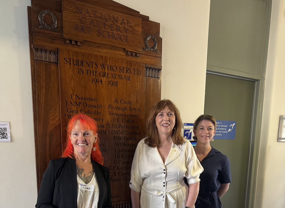 Three people stand in front of a wooden honour board