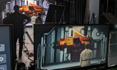 People stand on a film set in front of an orange spacecraft which also appears on a monitor screen