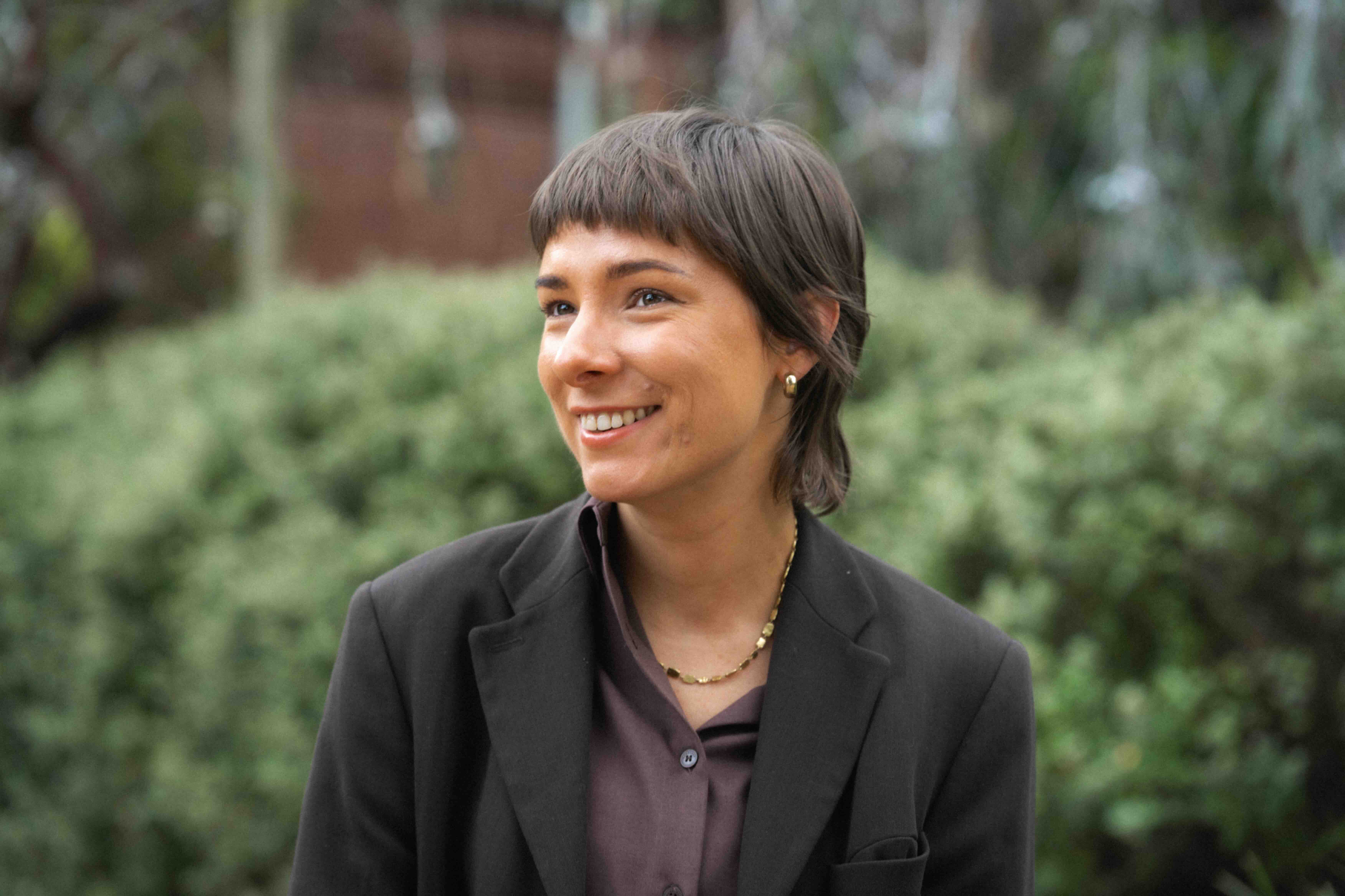 A person in a black suit stands in front of trees smiling