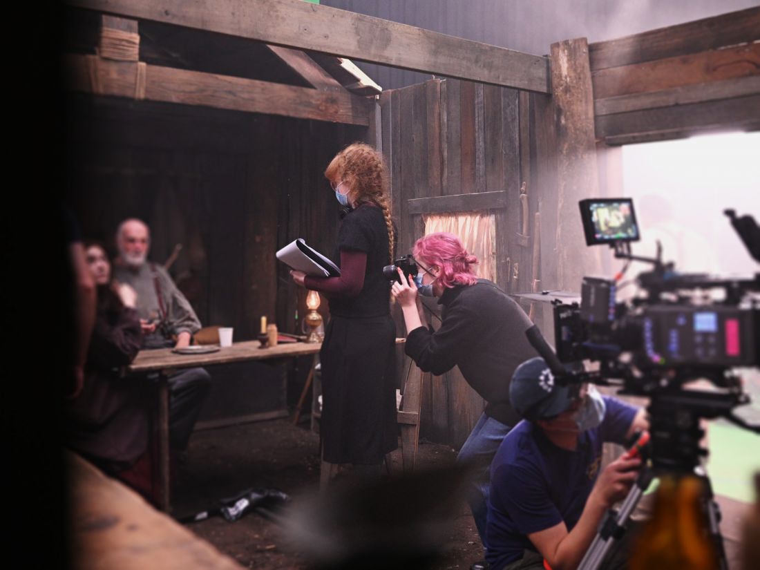 Sarah Hegge-Taylor stands in a wooden settler-style cabin film set, talking to actors seated at a table.