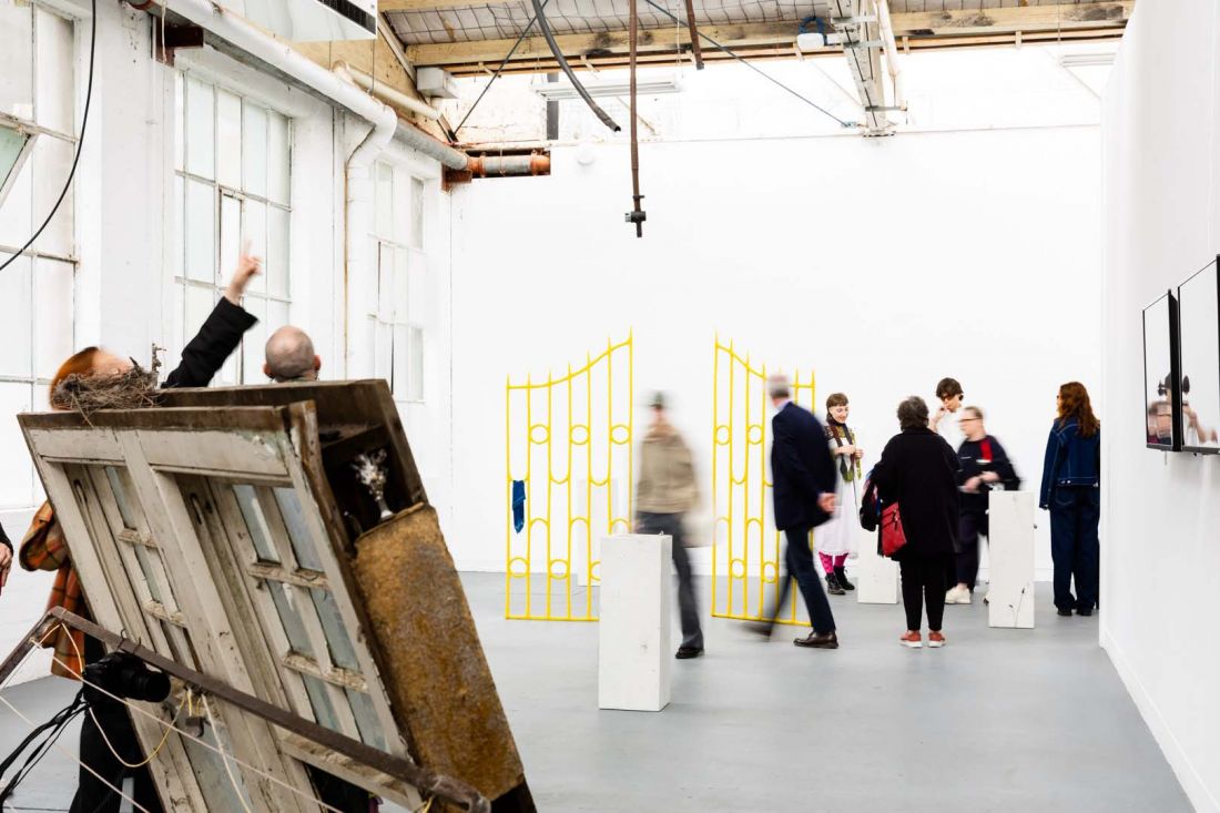 Brightly lit gallery space full of people filled with sculptural works, including a bright yellow sculptural gate