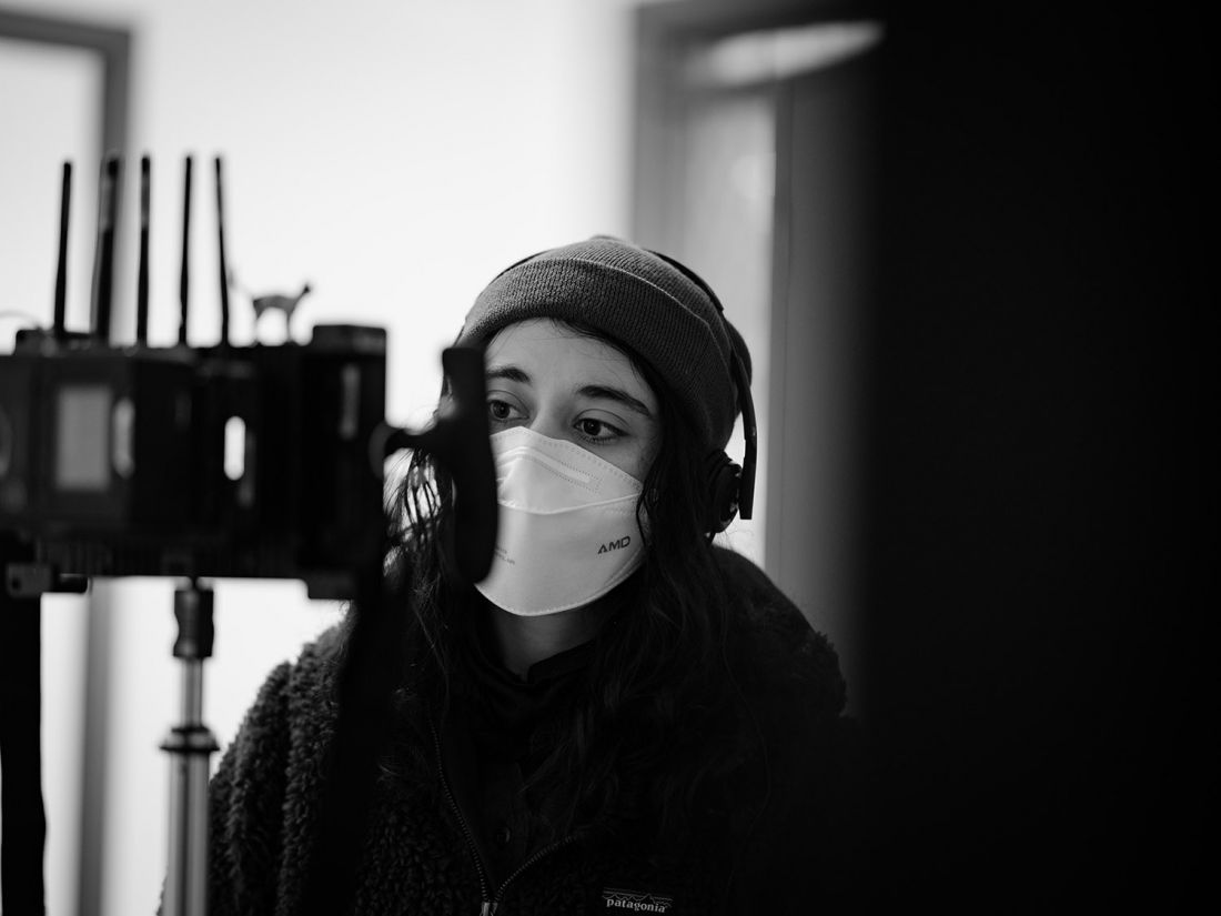Noora Niasari on set in black and white, sitting behind filming gear with headphone and mask on.
