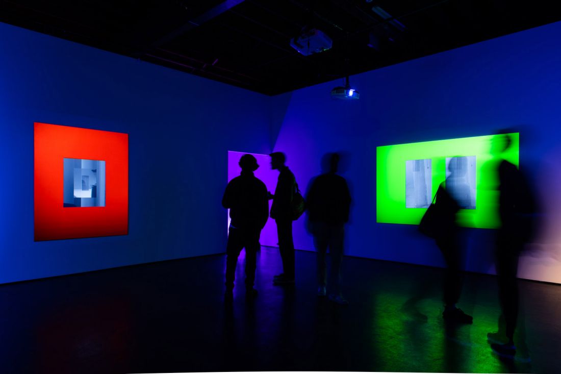 Darkly lit gallery space with silhouettes of people viewing glowing red and green artworks