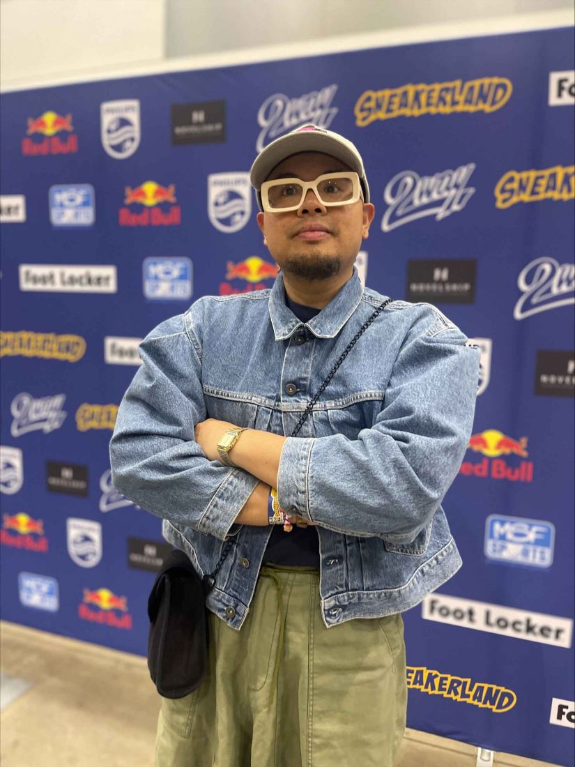 Victor standing in front of a logo wall wearing a denim jacket, a hat and glasses.