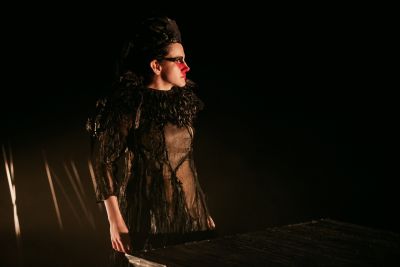 A woman stands on a dark stage in a costume of black feathers.